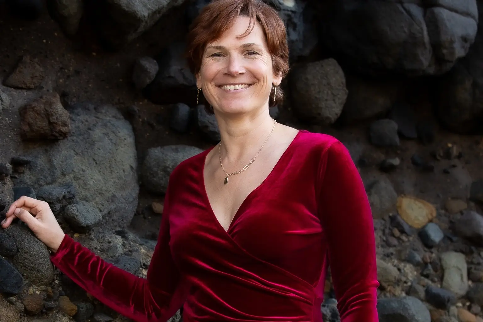 Tarisha Tourok in a red velvet dress, smiling by dark coastal rocks